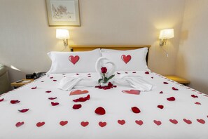 Un lit bien fait, décoré de pétales de roses et d’ornements en forme de cœur, deux lampes fixées au mur et un tableau encadré accroché au mur.