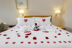 Una cama bien hecha con pétalos de rosa y adornos con forma de corazón, dos lámparas fijadas a la pared y un cuadro en la pared.