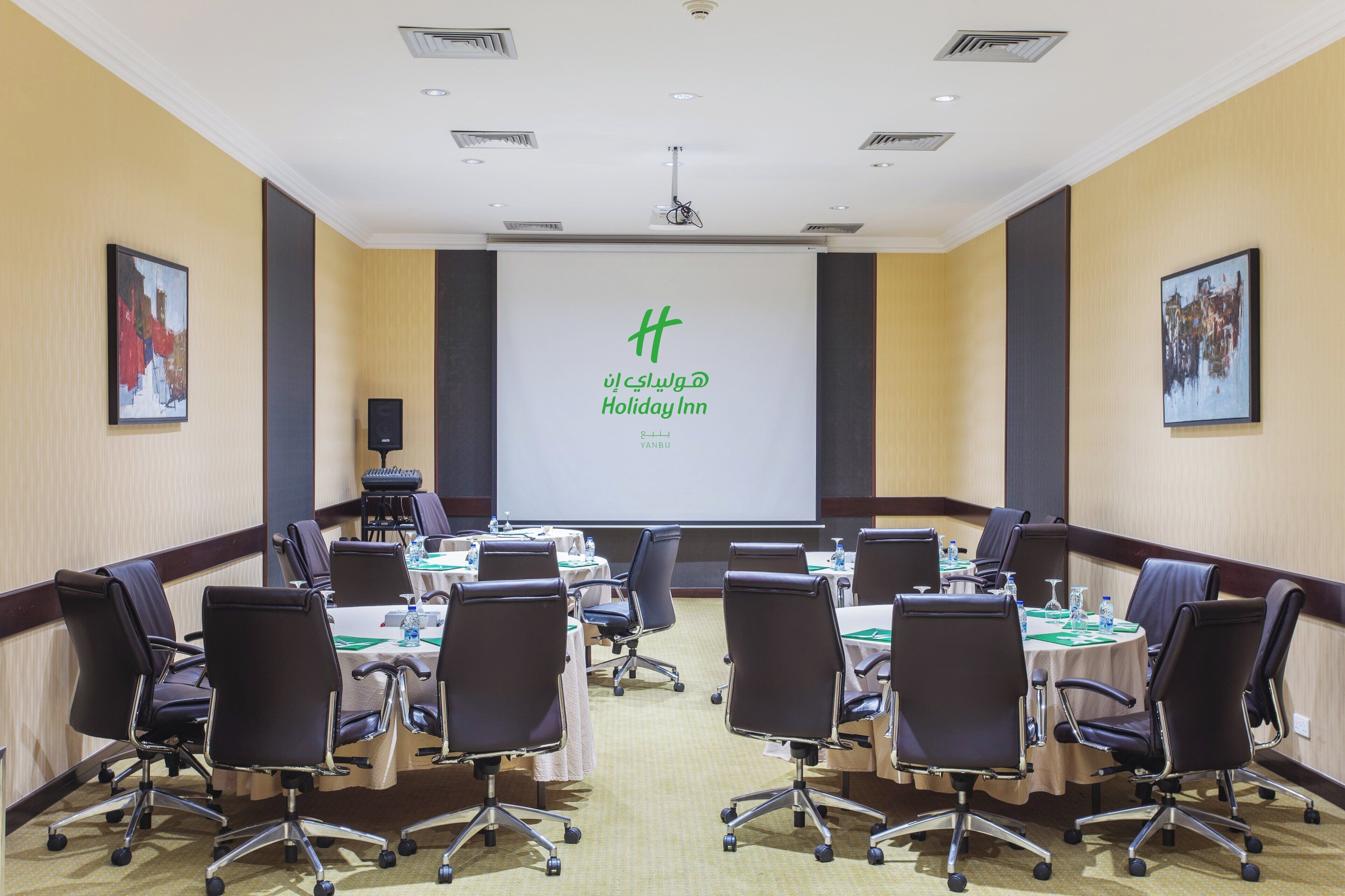 A conference room with a projector screen displaying 'Holiday Inn' branding, multiple tables with chairs, and a carpeted floor.