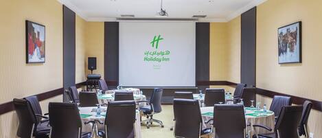 A conference room with a projector screen displaying 'Holiday Inn' branding, multiple tables with chairs, and a carpeted floor.