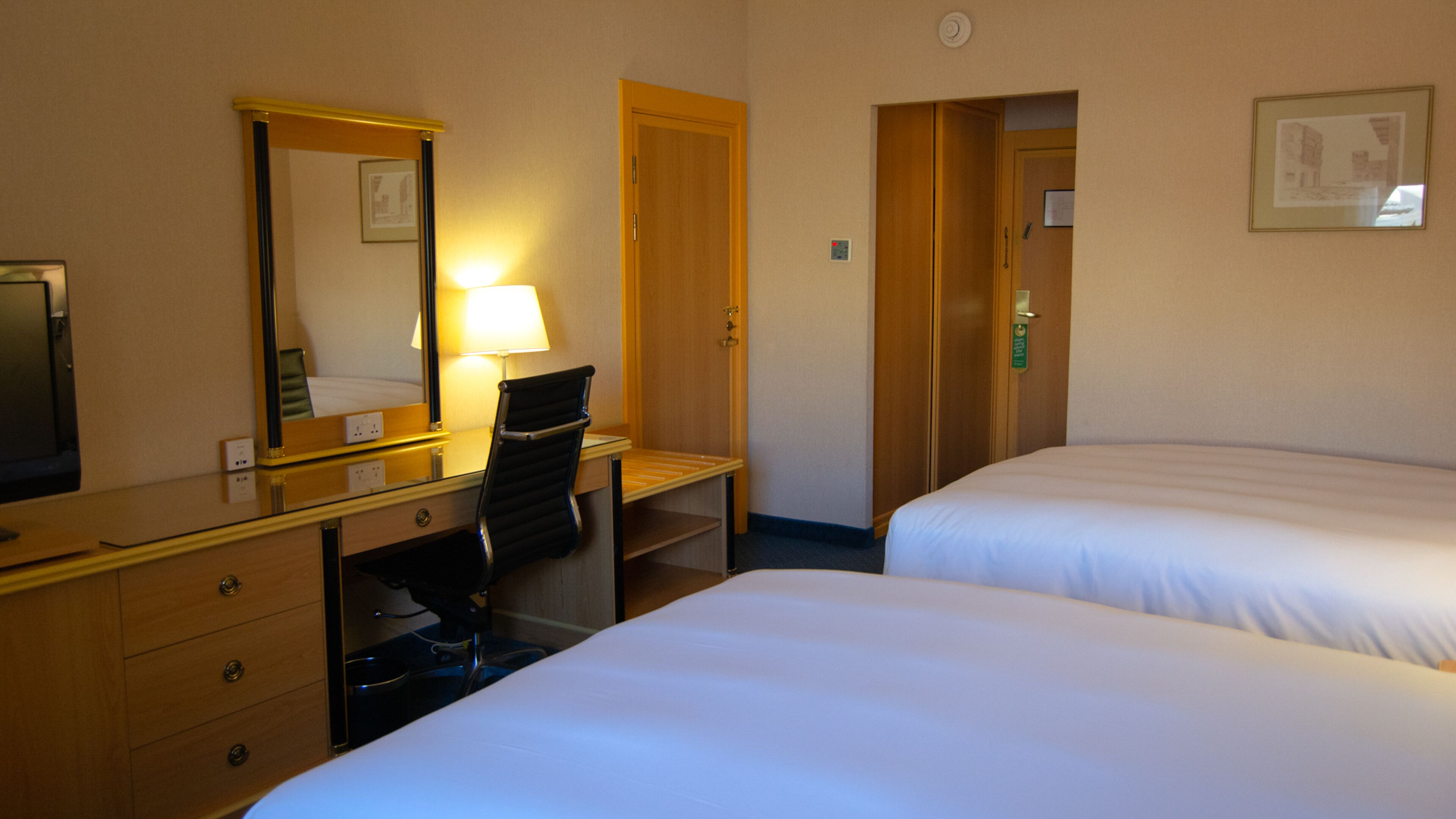 A hotel room with a desk, chair, two beds, a mirror, and a door.