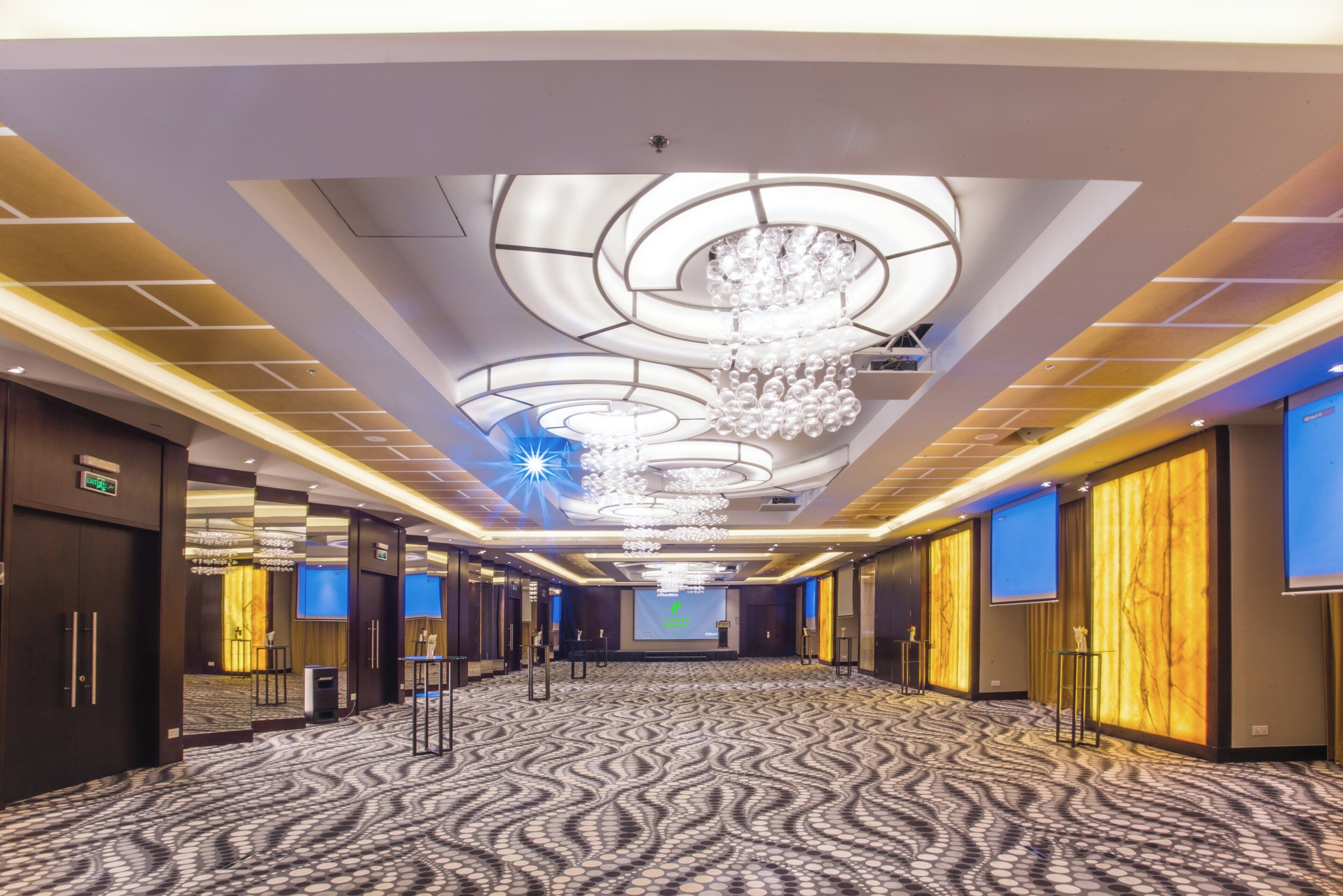 A hotel corridor with a patterned carpet, a large chandelier, and a projection screen at the end.