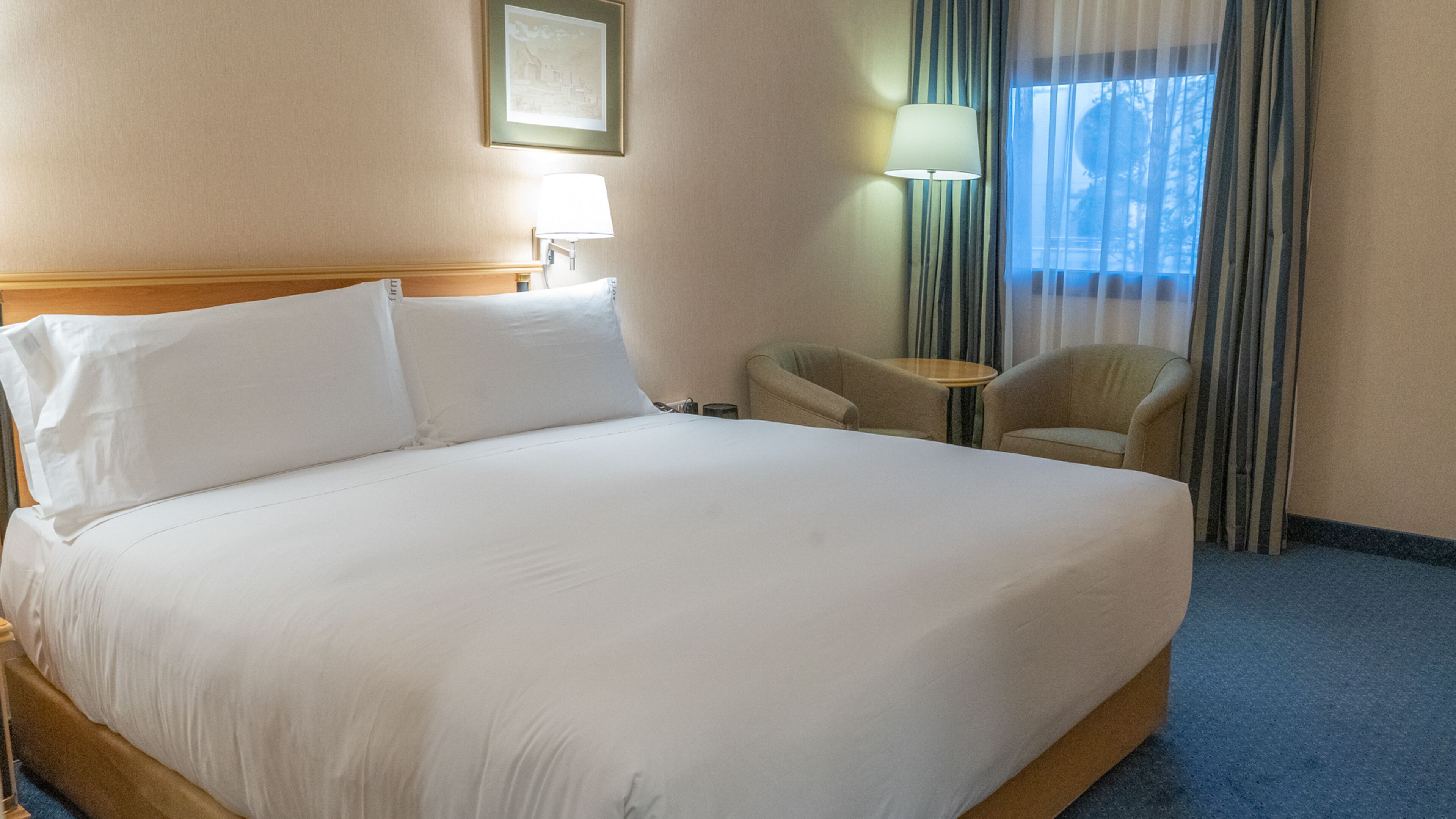 Een hotelkamer met een groot bed, een stoel, een klein tafeltje en een lamp.