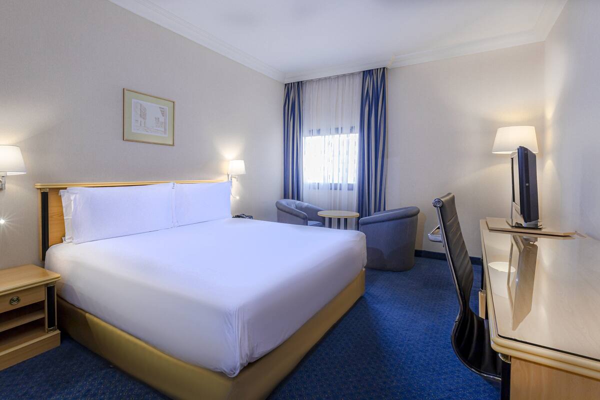 A hotel room with a large bed, bedside tables, a desk with a chair, a television, and a window with curtains.