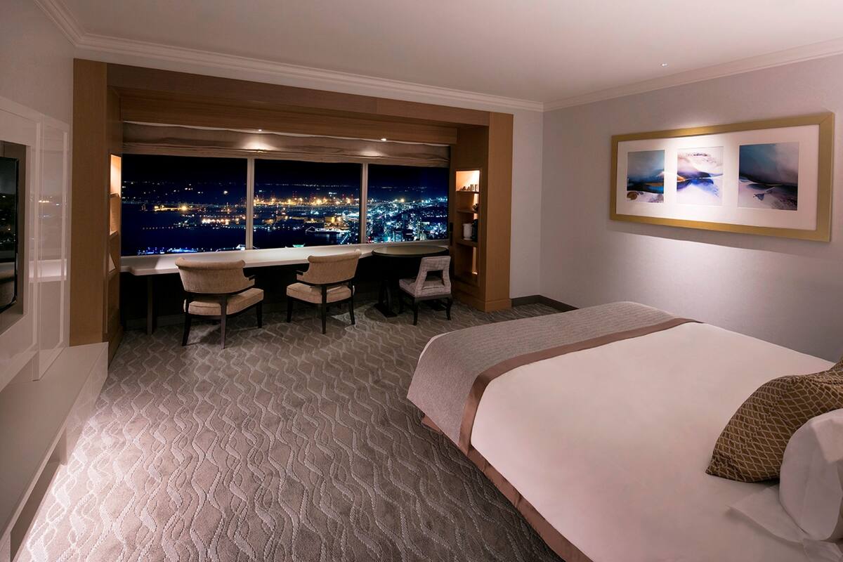 A hotel room with a large bed, a desk with two chairs, and a view of a cityscape at night.