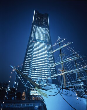 Un rascacielos alto e iluminado con un barco atracado al frente, destacando una proa grande y ornamentada y el nombre "NIPPON MARU".
