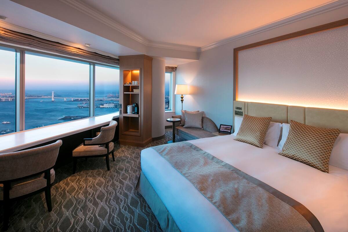 A hotel room with a large bed, a desk with chairs, a small table with a lamp, and a view of the city and bridge.