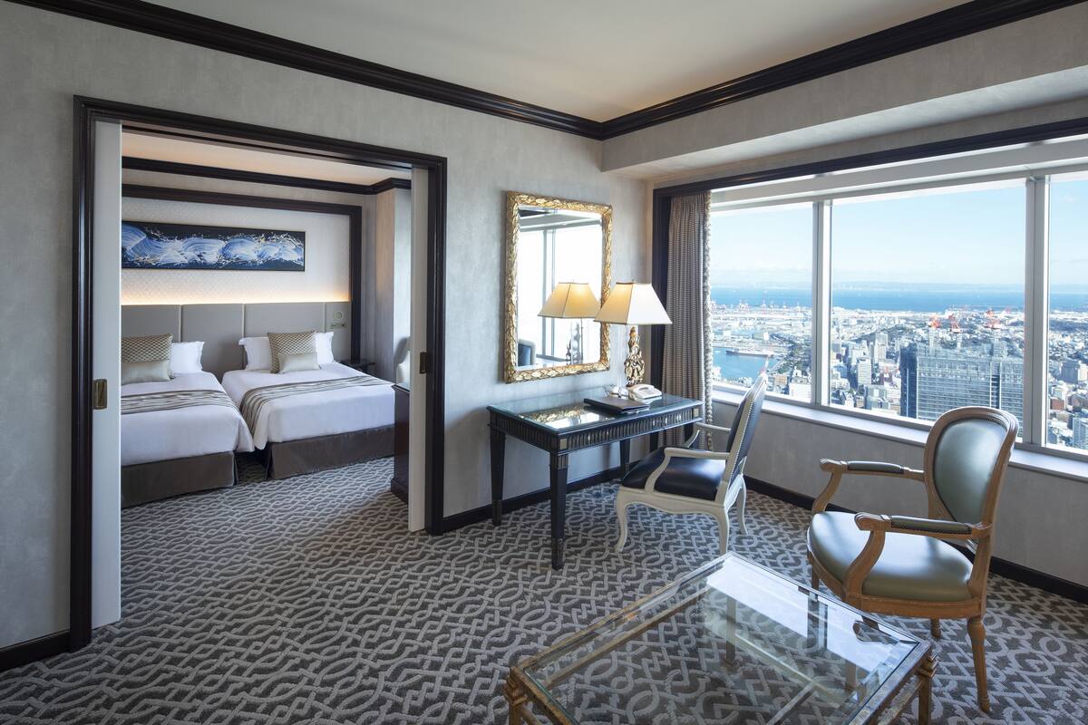 A hotel room with a bed, a desk with a chair, a large window with a city view, and a patterned carpet.