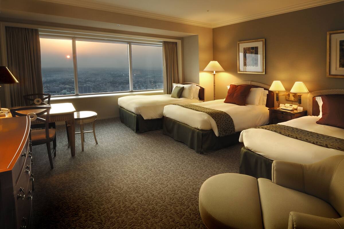 A hotel room with two beds, a desk, a chair, and a large window with a city view.