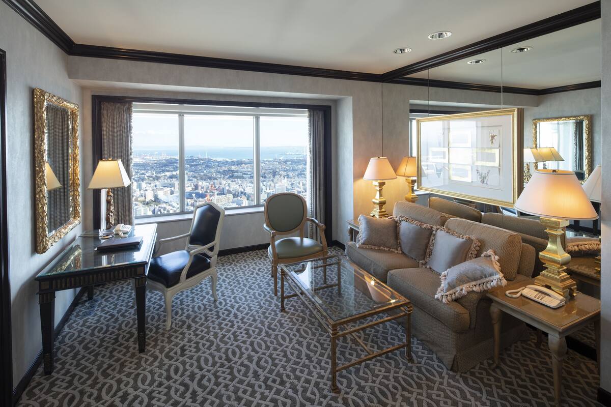 A hotel room with a city view, a desk with a lamp, a sofa, and a chair.