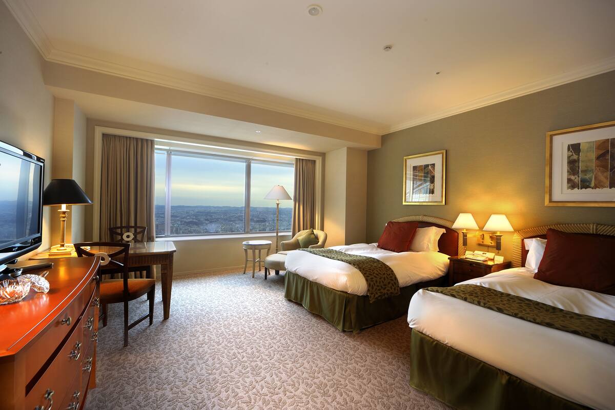 A hotel room with two beds, a desk, a TV, and a large window offering a city view.