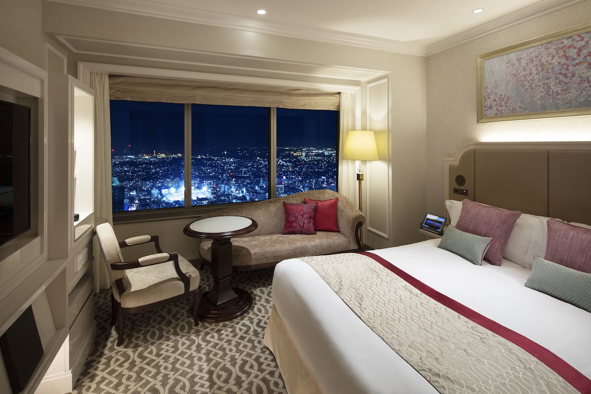 A hotel room with a large bed, a sofa, a small table, and a view of a cityscape at night.