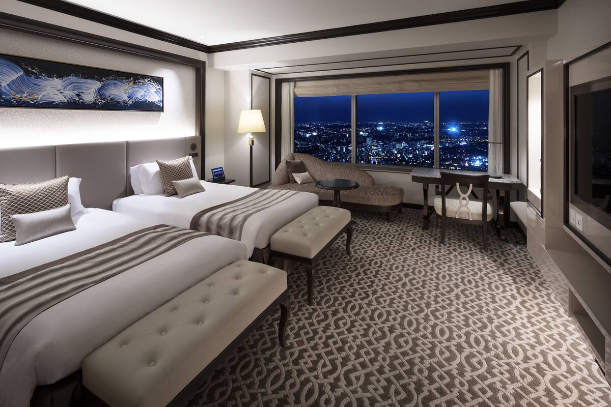 A hotel room with two beds, a small table, a chair, and a view of the city at night.