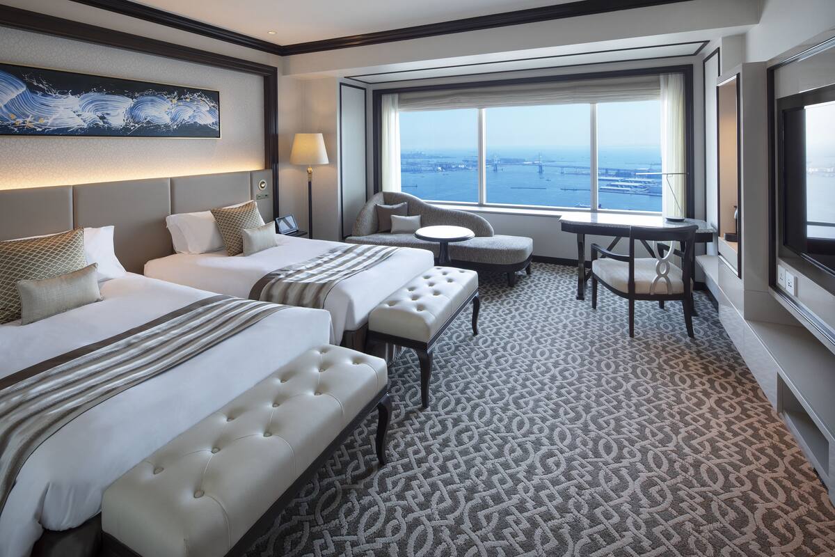 A hotel room with two beds, a sofa, a small table, and a view of the cityscape.