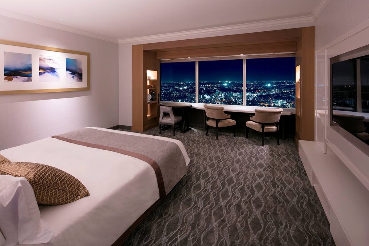 A hotel room with a bed, a desk, two chairs, a large window with a city view, and a framed picture on the wall.