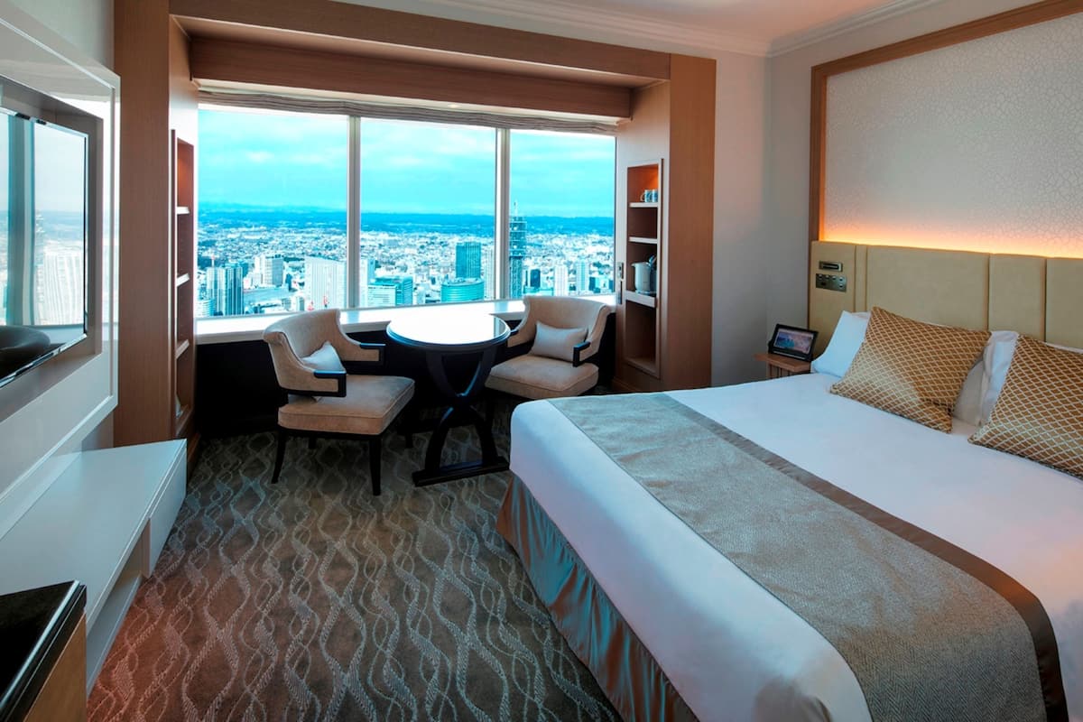 A hotel room with a large bed, a desk with two chairs, a view of a cityscape through large windows, and a bookshelf.