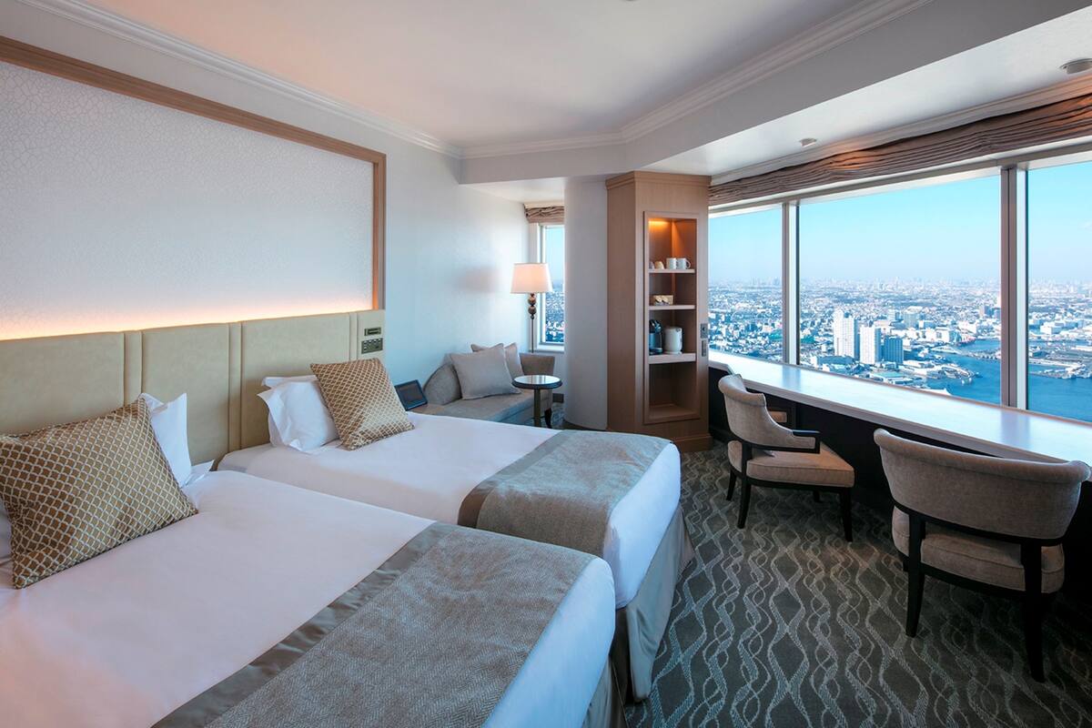 A hotel room with two beds, a desk, and a view of the city.