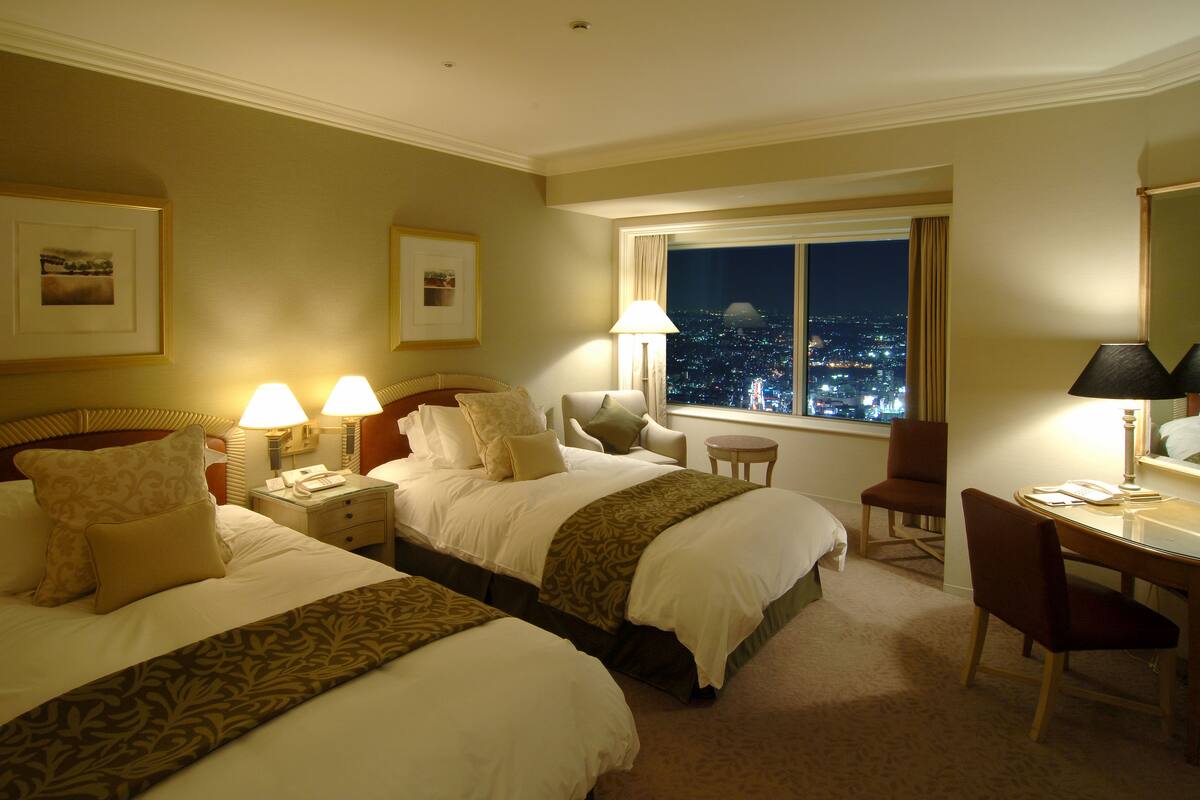 A hotel room with two beds, a desk, a chair, and a window with a city view at night.