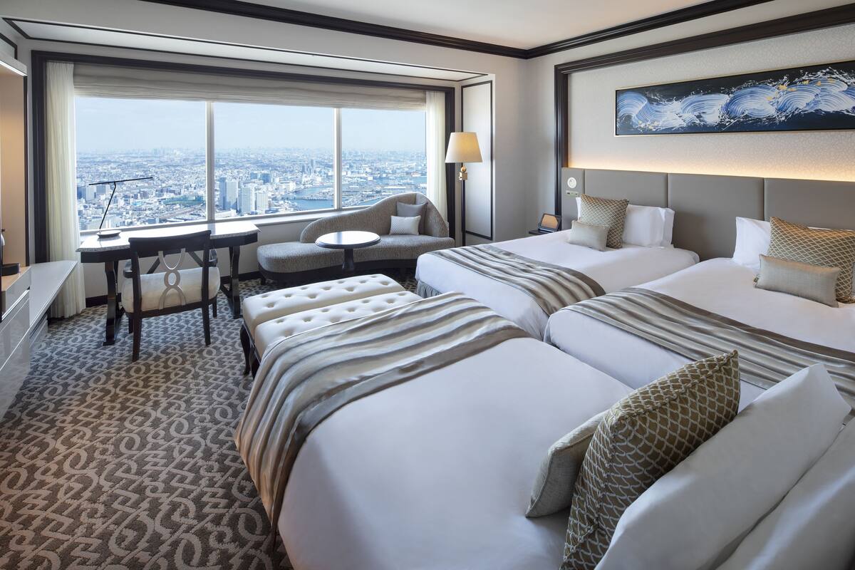 A hotel room with two beds, a desk, a chair, and a large window offering a city view.