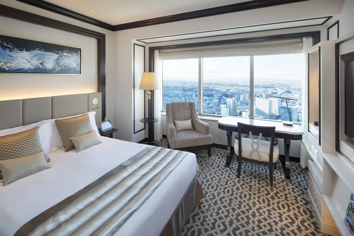 A hotel room with a large bed, a desk with a chair, a view of the city, and a painting on the wall.