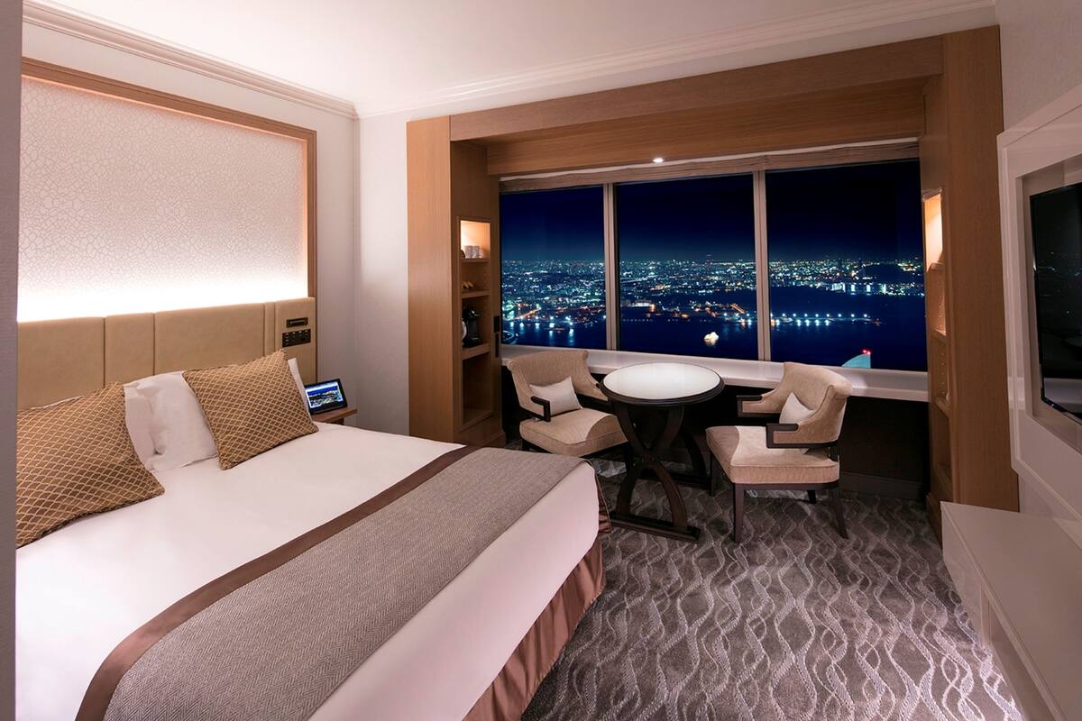 A hotel room with a large bed, a desk, two chairs, a view of a cityscape at night, and a television on the wall.