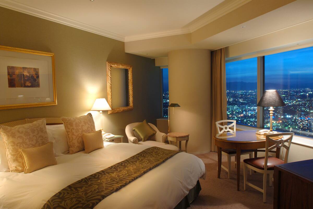 A hotel room with a large bed, a desk, and a chair. There is a view of a cityscape through the window.