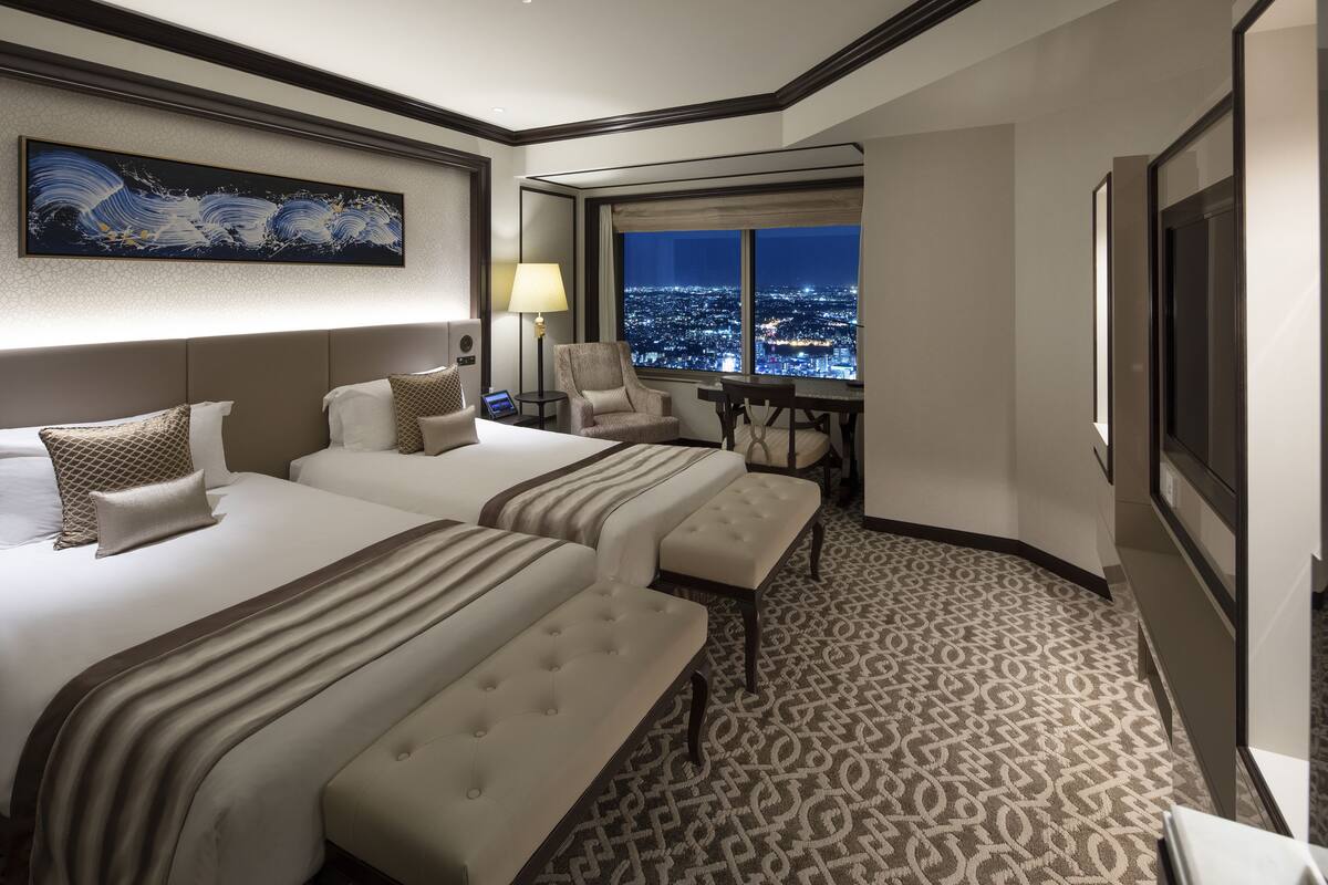 A hotel room with two beds, a desk, a chair, a large window with a city view, and a painting on the wall.