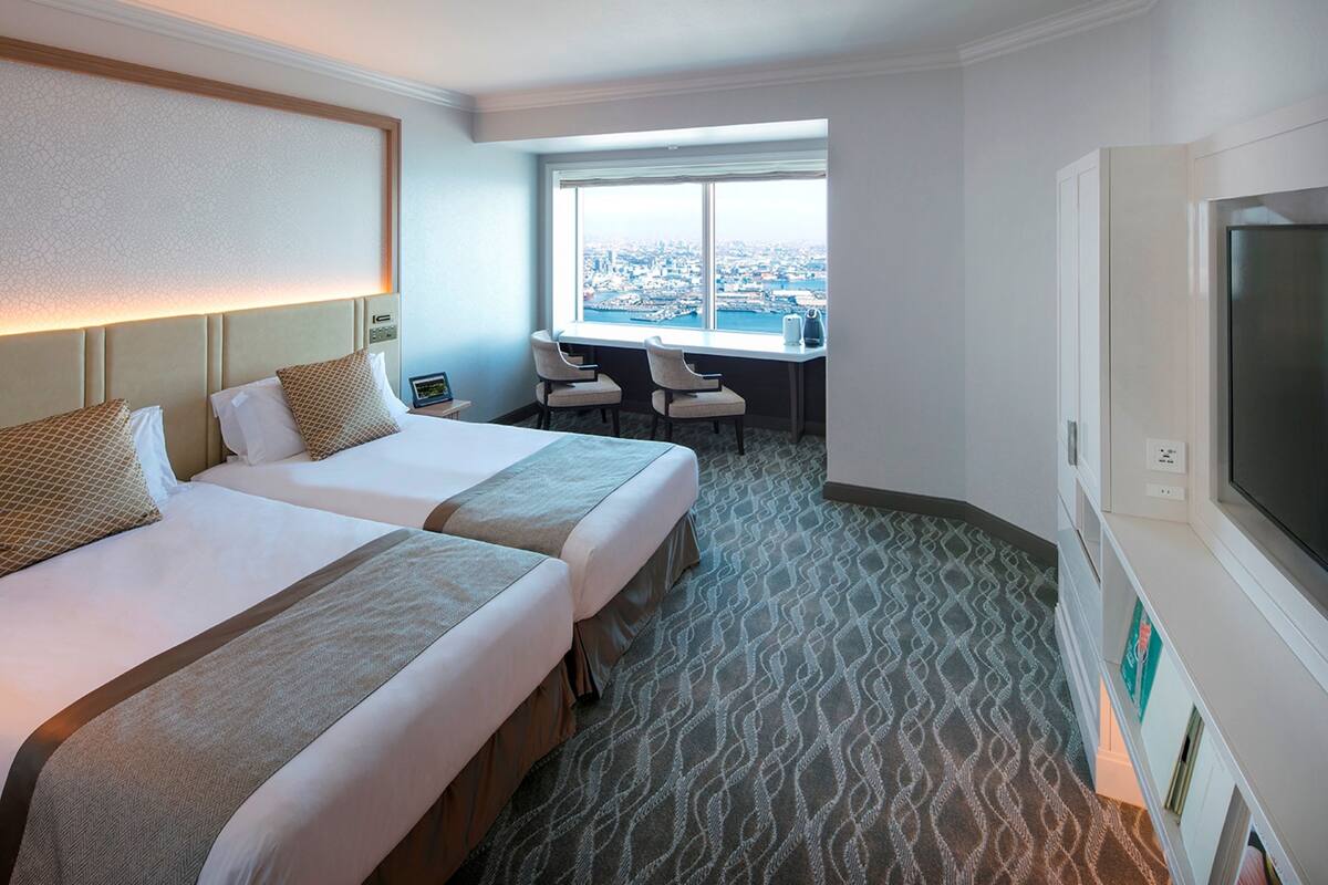 A hotel room with two beds, a desk with chairs, a large window with a city view, and a TV.