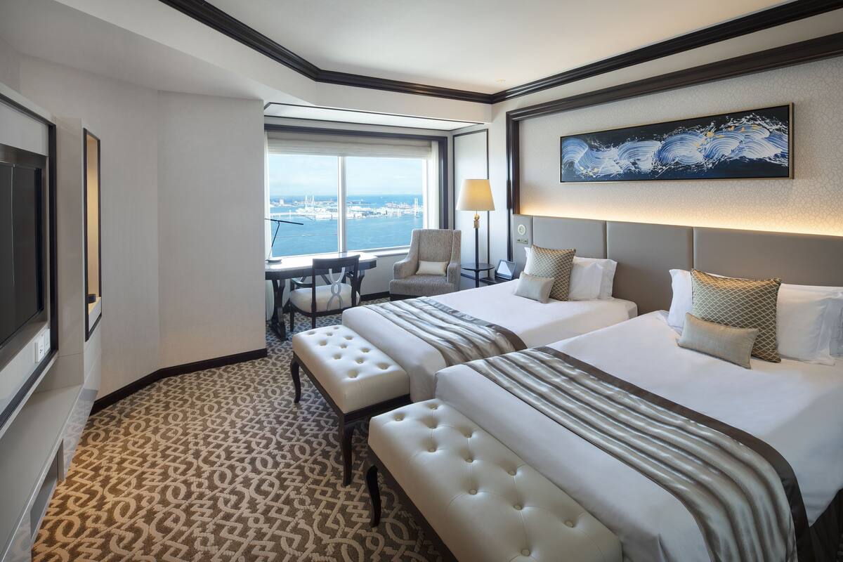A hotel room with two beds, a large window with a city view, a desk with a chair, and a lamp.