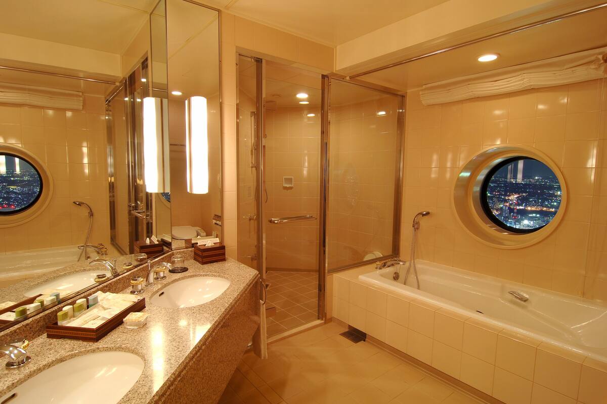 A modern bathroom with two sinks, a glass-enclosed shower, and a bathtub with a city view window.