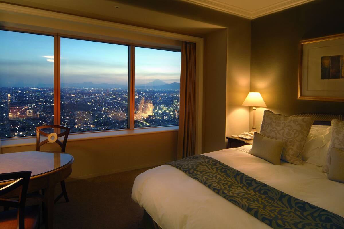 A hotel room with a large window offering a city view, a bed with patterned bedding, a bedside table with a lamp, and a chair near the window.