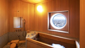 Un baño de madera con una bañera grande, un taburete pequeño de madera, un espejo y una ventana con vistas a la ciudad.