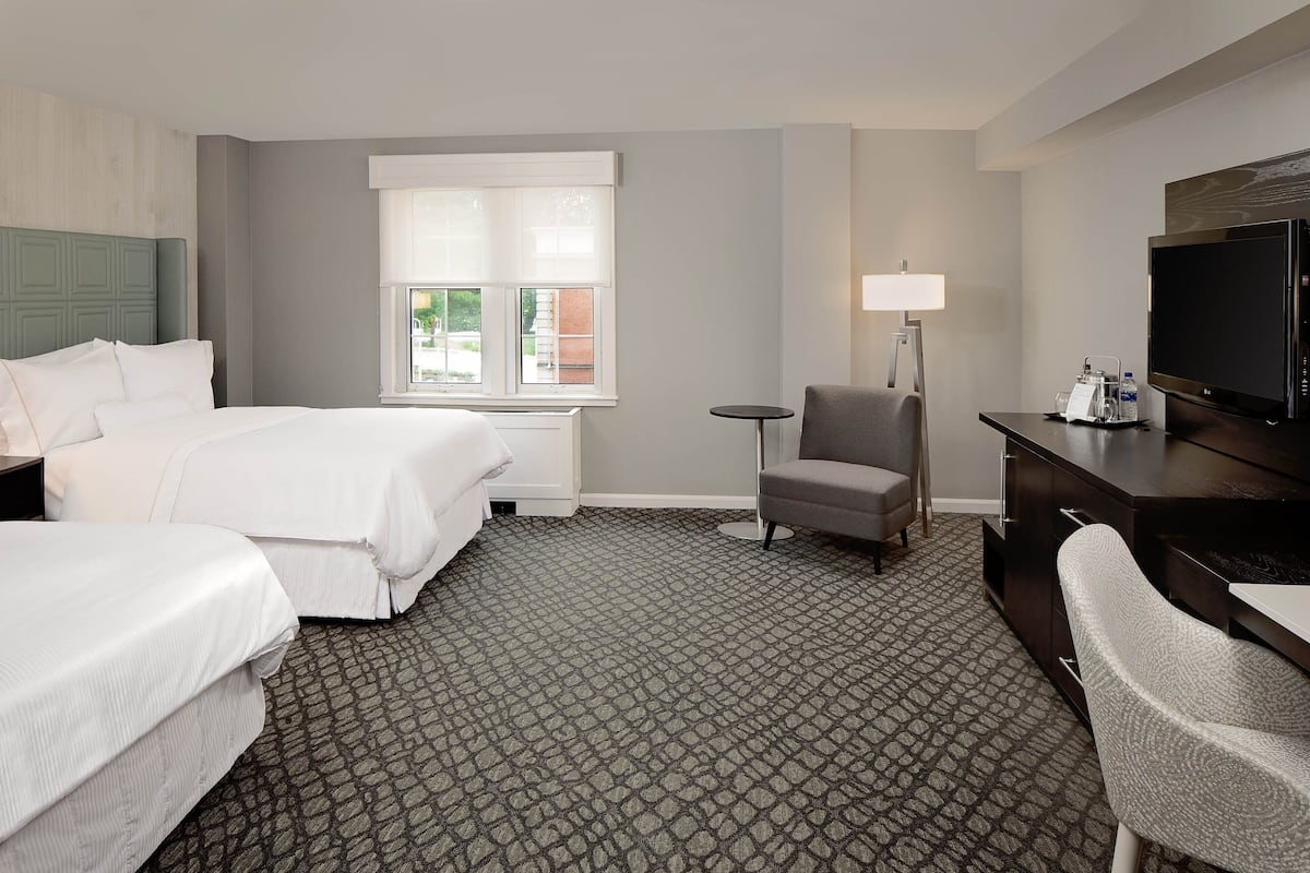 A hotel room with two beds, a desk, a chair, a television, and a lamp.