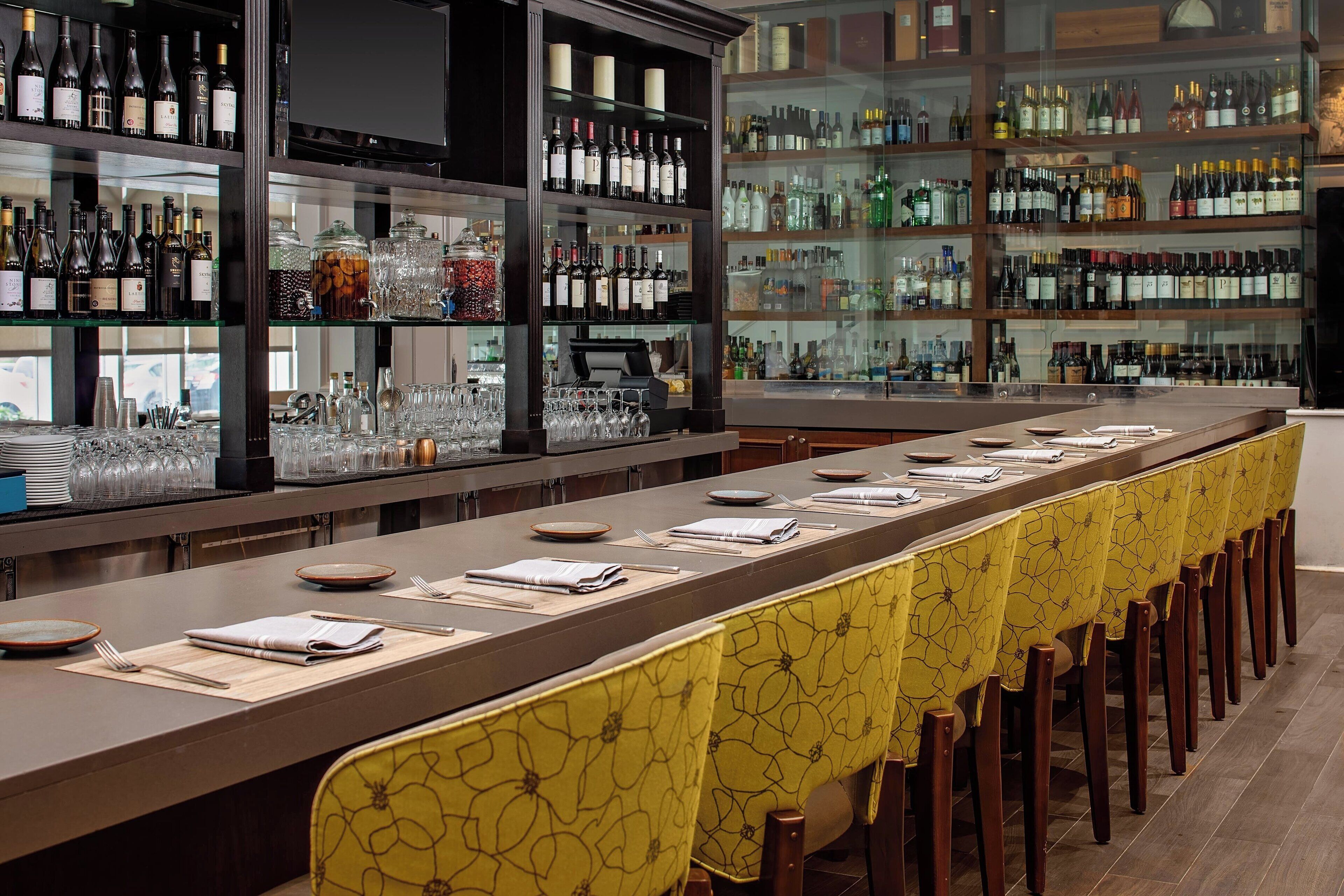 A long bar with neatly arranged glasses and bottles, a long table with chairs covered in yellow fabric, and shelves stocked with various bottles and glasses.