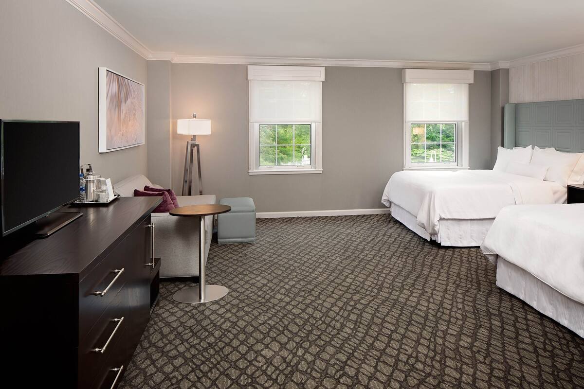 A hotel room with two beds, a desk, a TV, and a window with a view of greenery.