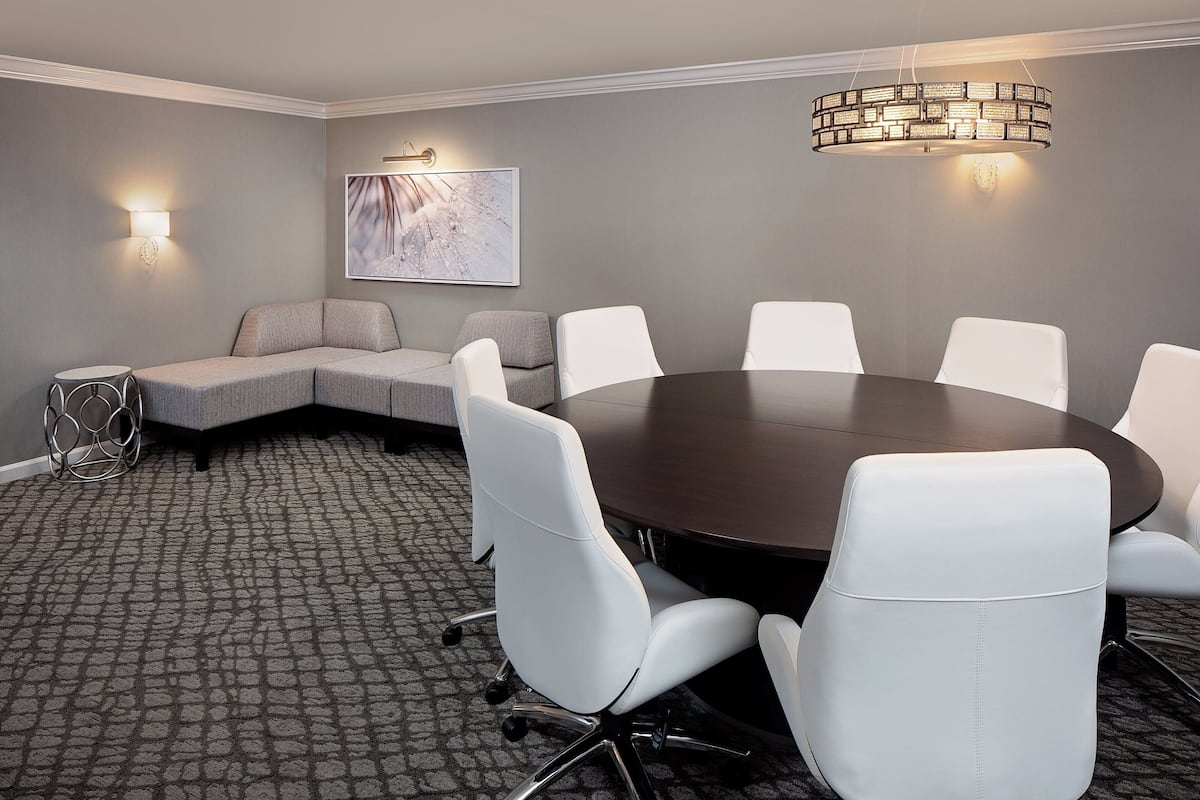 A conference room with a round table, white chairs, a gray sofa, a patterned carpet, and a wall-mounted light fixture.
