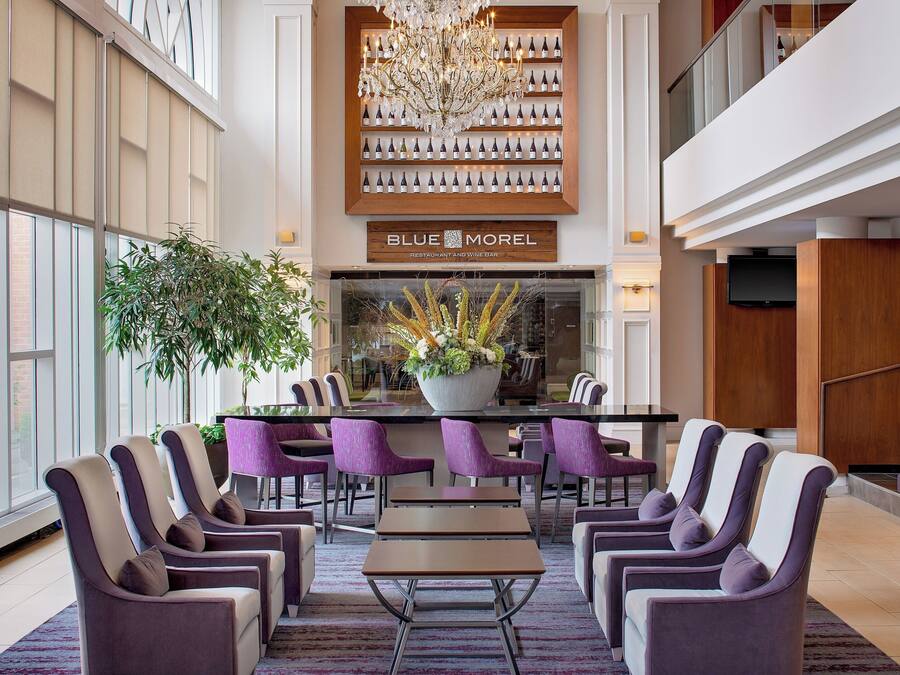 A modern hotel lobby with a reception desk, purple chairs, a chandelier, and a large window.