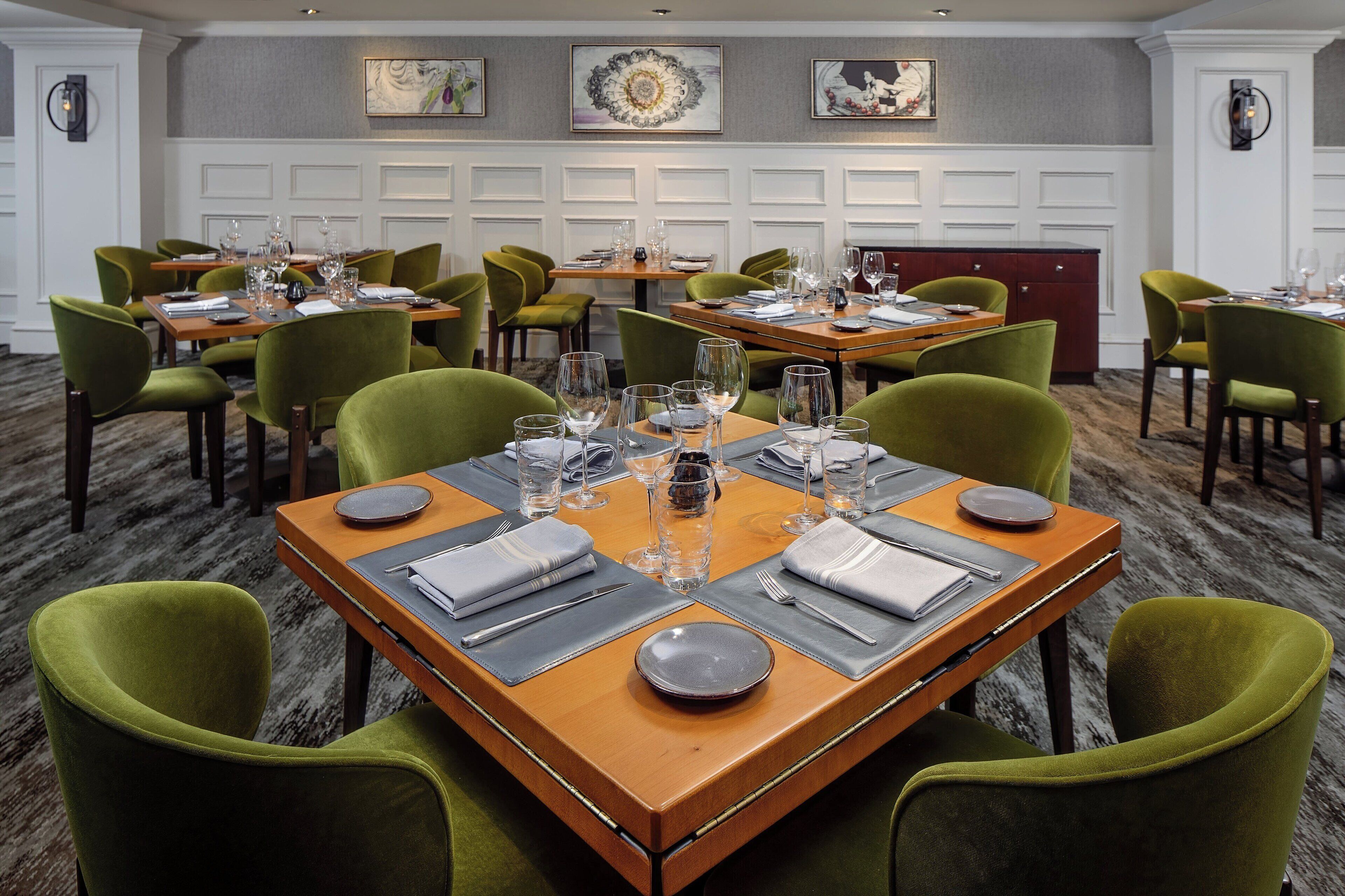 Un salón de conferencias con varias mesas dispuestas para comidas, cada una con sillas tapizadas en verde, manteles blancos y cubertería y cristalería ordenadas.
