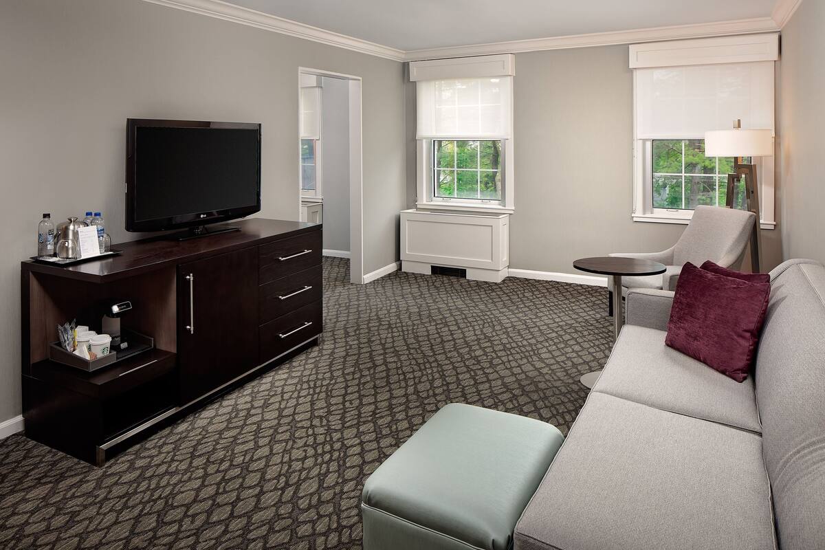 A hotel room with a dark wooden dresser, a flat-screen TV, a grey sofa, and a small round table.