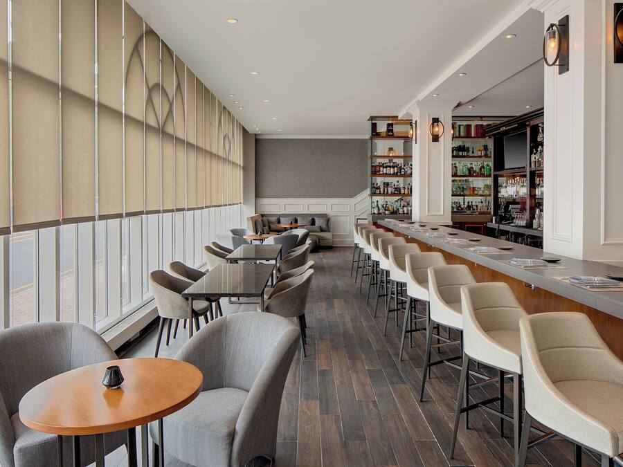 Una zona de bar moderna con una barra alargada, diversas opciones de asientos, incluyendo taburetes y mesas, y una amplia estantería para vinos al fondo.