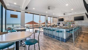 Eine Bar mit Blick auf den Ozean, Tische, Stühle und eine Bar, die mit verschiedenen Flaschen bestückt ist.