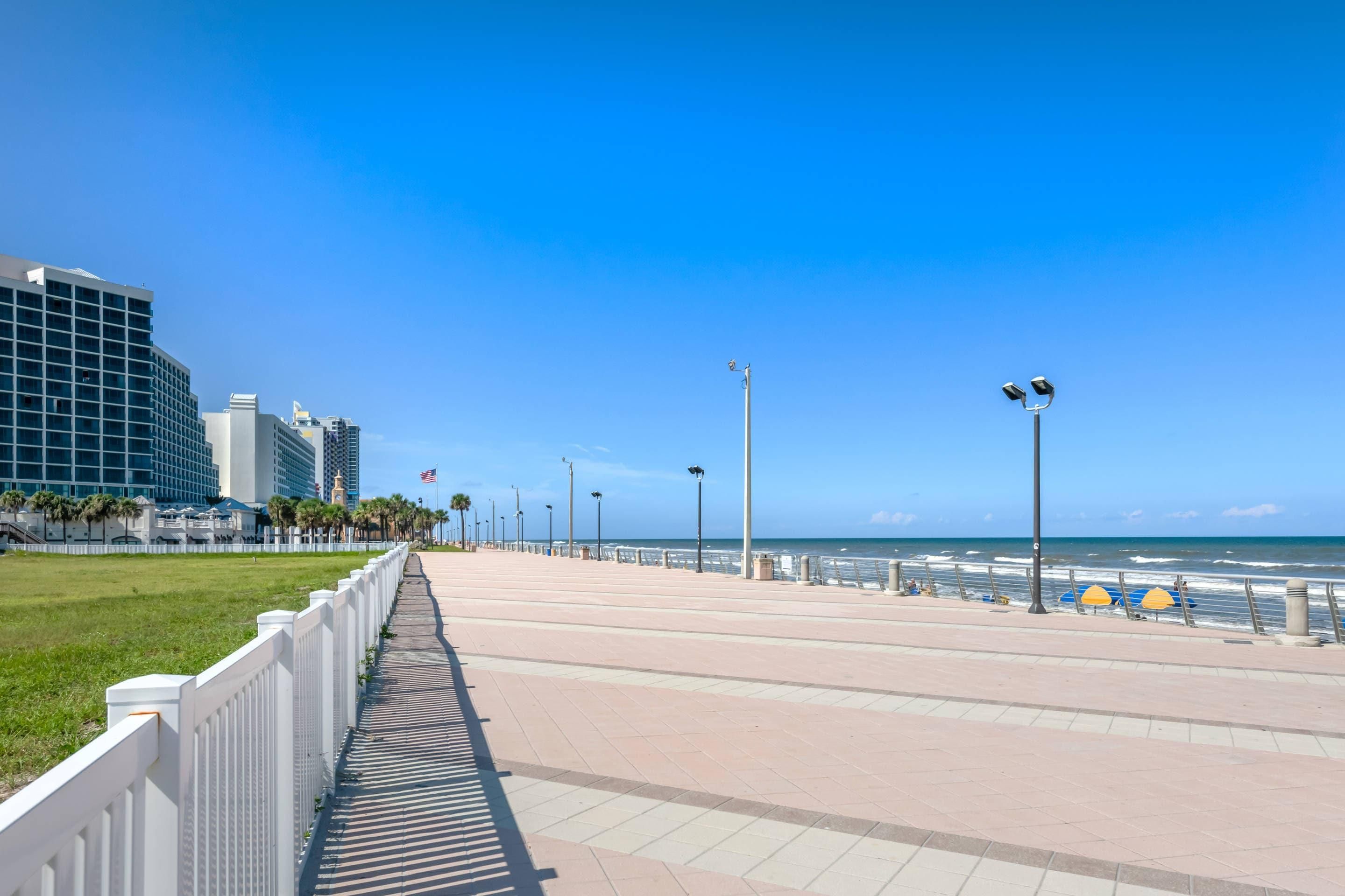 Un paseo marítimo con un camino pavimentado, cerca de una valla blanca, farolas y vistas al océano.