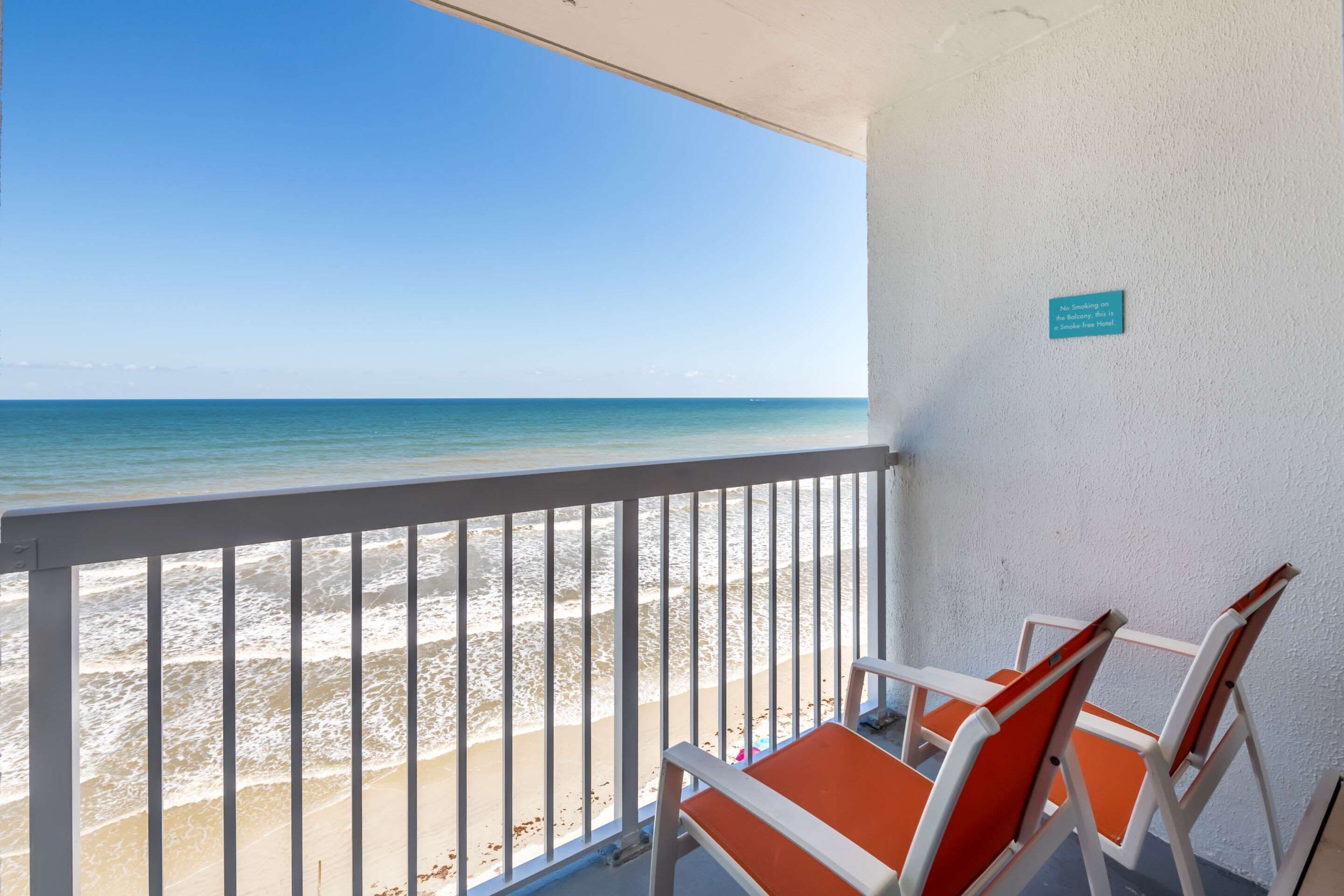 A balcony with a view of the beach and ocean. There are two orange and white chairs. A sign on the wall reads "No Smoking on the Balcony Area."