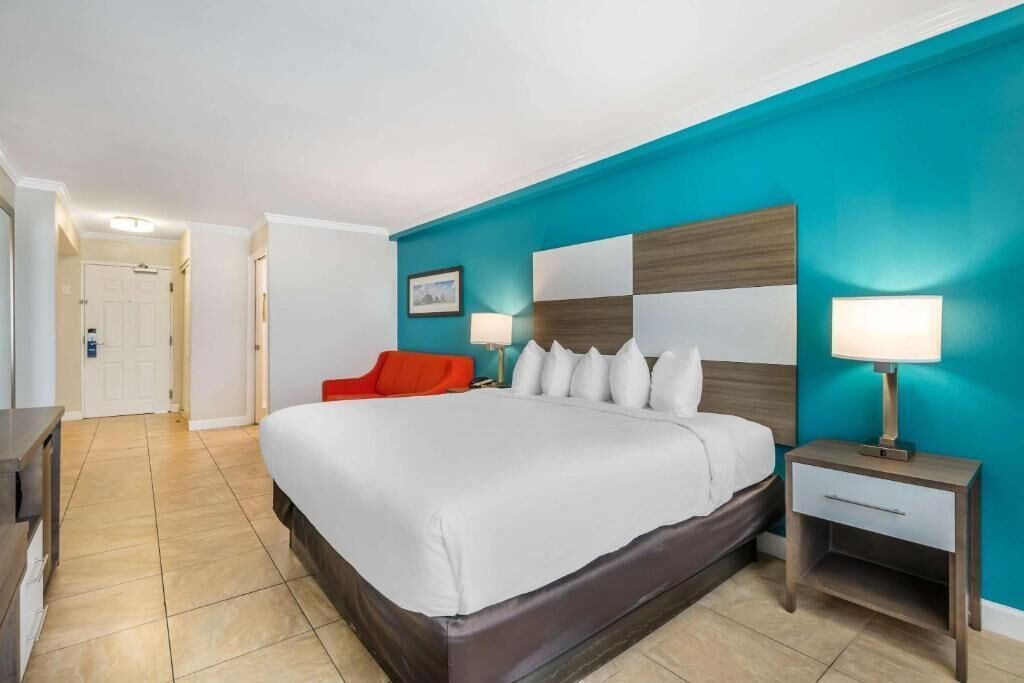 A hotel room with a large bed, a red chair, a nightstand with a lamp, and a wall with a checkered pattern.