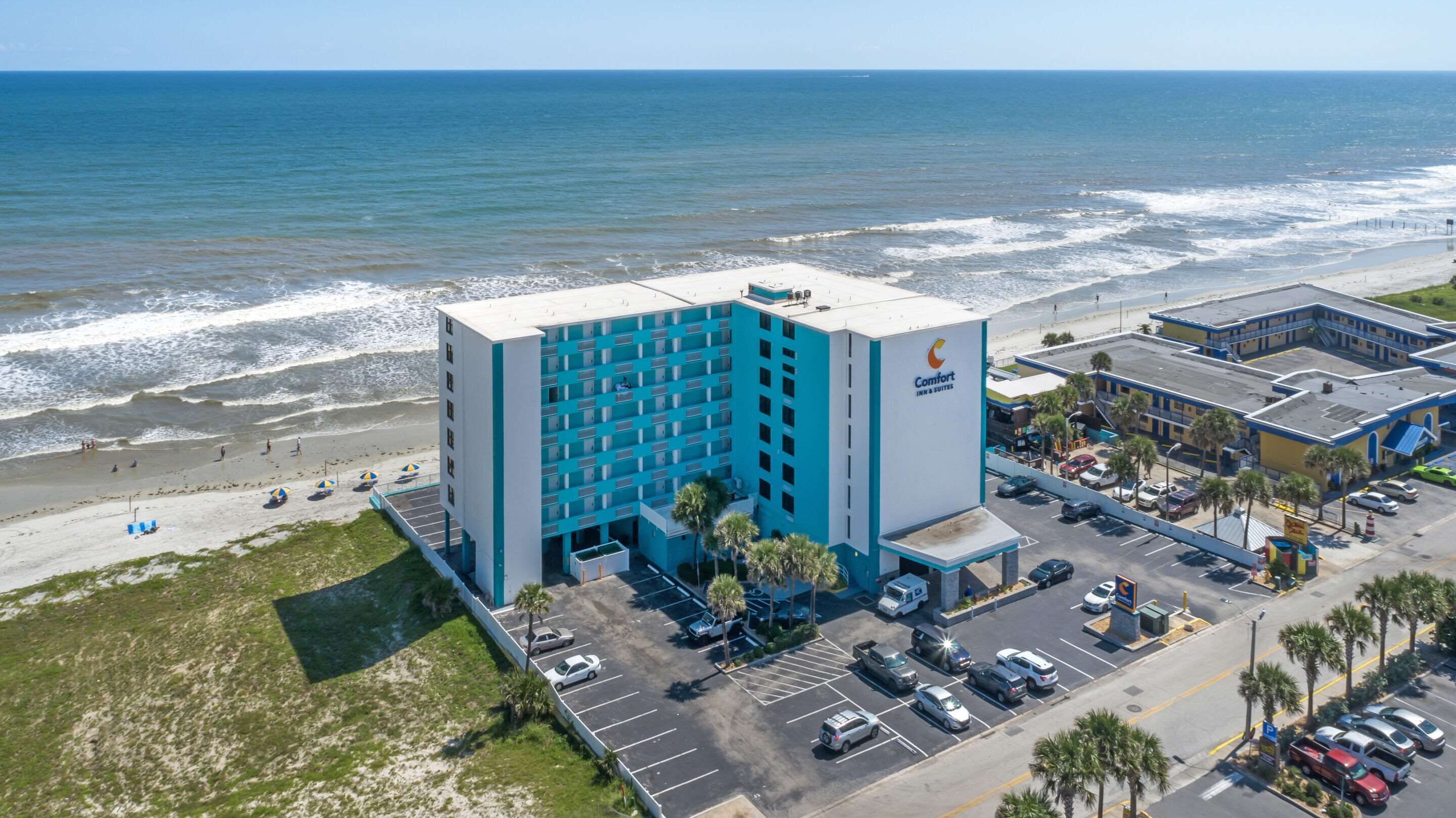 Un hotel con fachada azul y blanca, cerca de una playa que cuenta con aparcamiento y una carretera.
