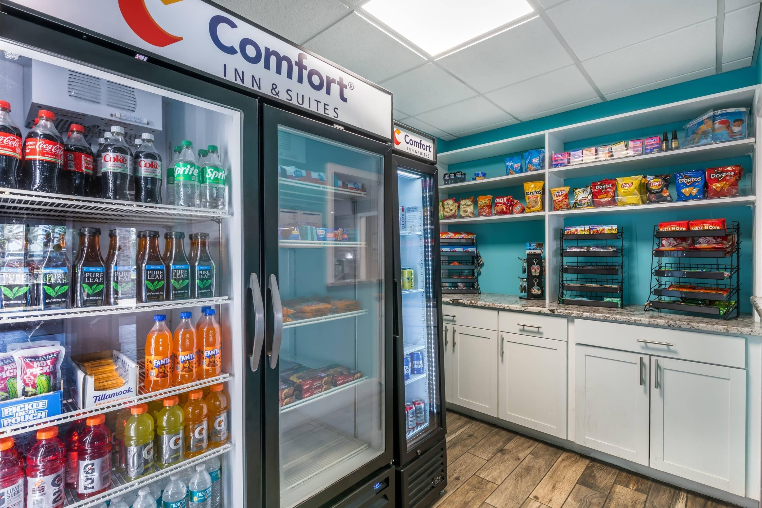 Una kitchenette de hotel con un refrigerador lleno de bebidas, un estante con bocadillos y una encimera con una máquina de café.