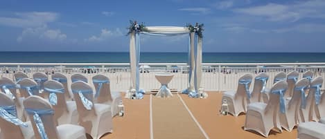 A beachside wedding setup with rows of white chairs and a central aisle leading to an arch decorated with greenery and flowers.