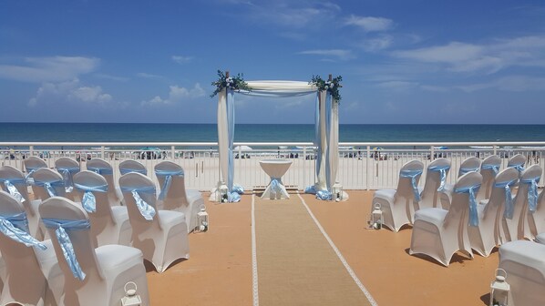 A beachside wedding setup with rows of white chairs and a central aisle leading to an arch decorated with greenery and flowers. - Comfort Inn & Suites Daytona Beach Oceanfront (Daytona Beach)
