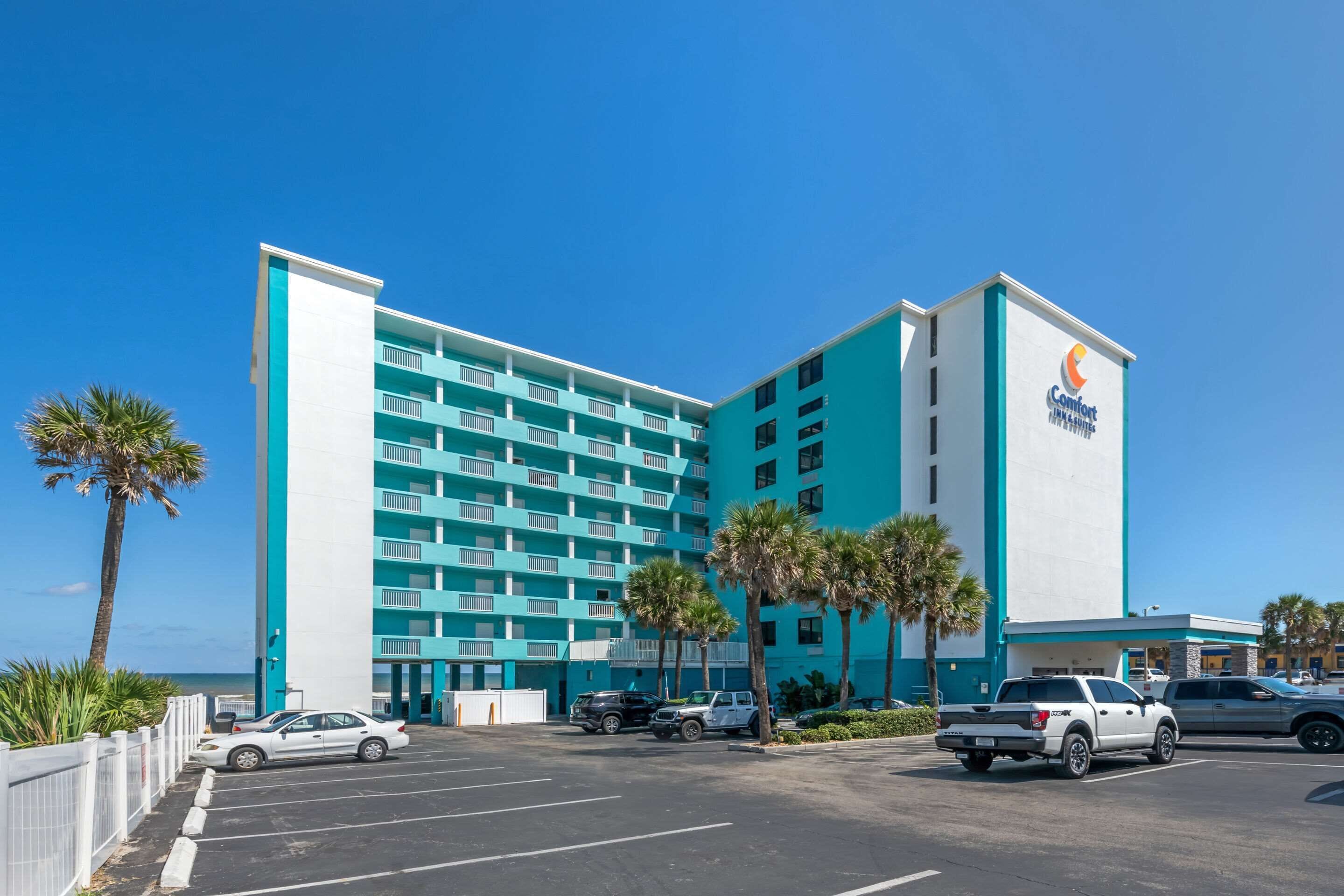 Een meerlaags hotel met een blauw-wit uiterlijk, een parkeerplaats en palmbomen.