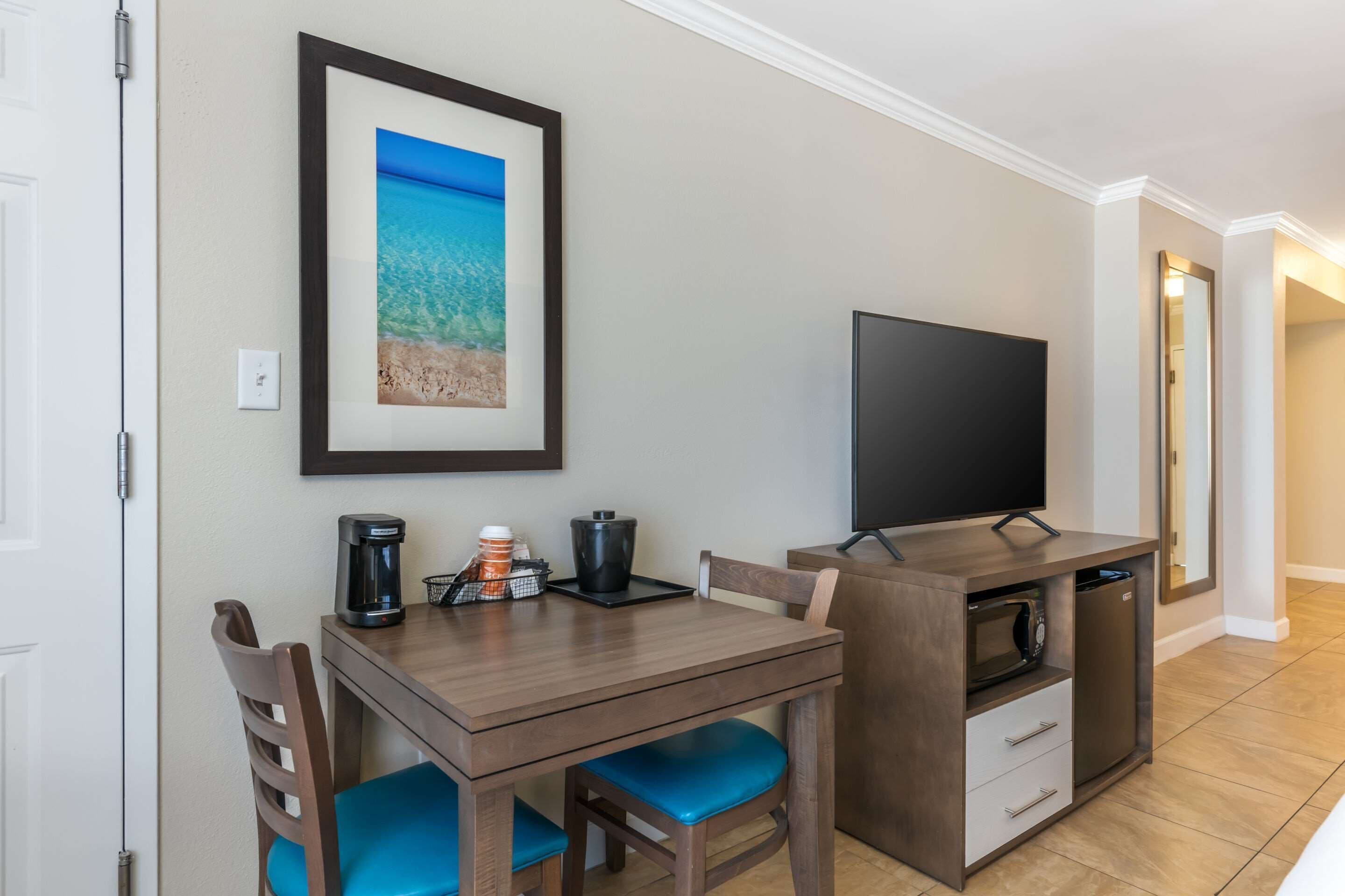 A compact room with a wooden table, blue cushioned chairs, a flat-screen TV, a coffee maker, and a framed picture of a beach scene.